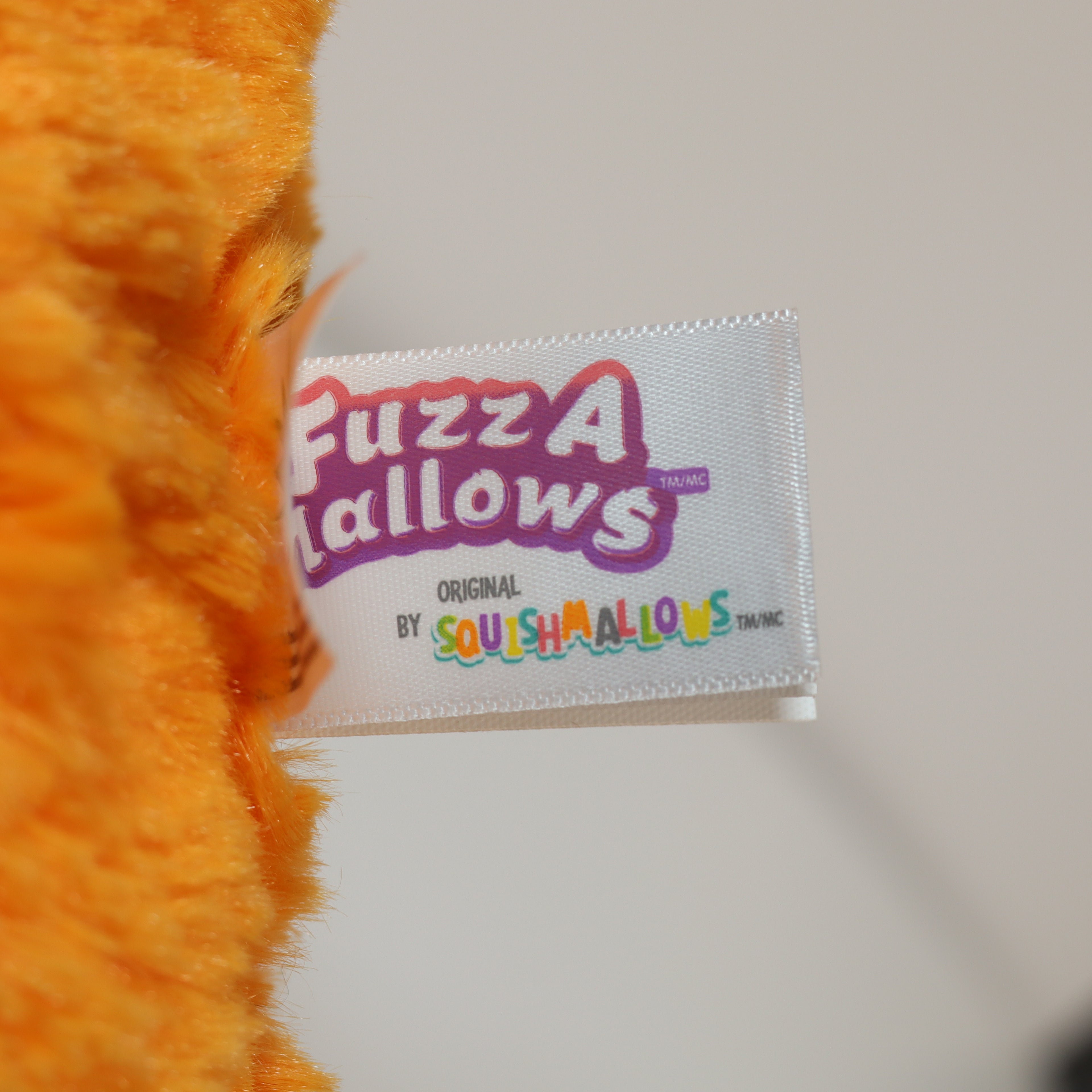 Close-up of an orange plush toy with a 'Fuzz A Mallovs' label on a white background
