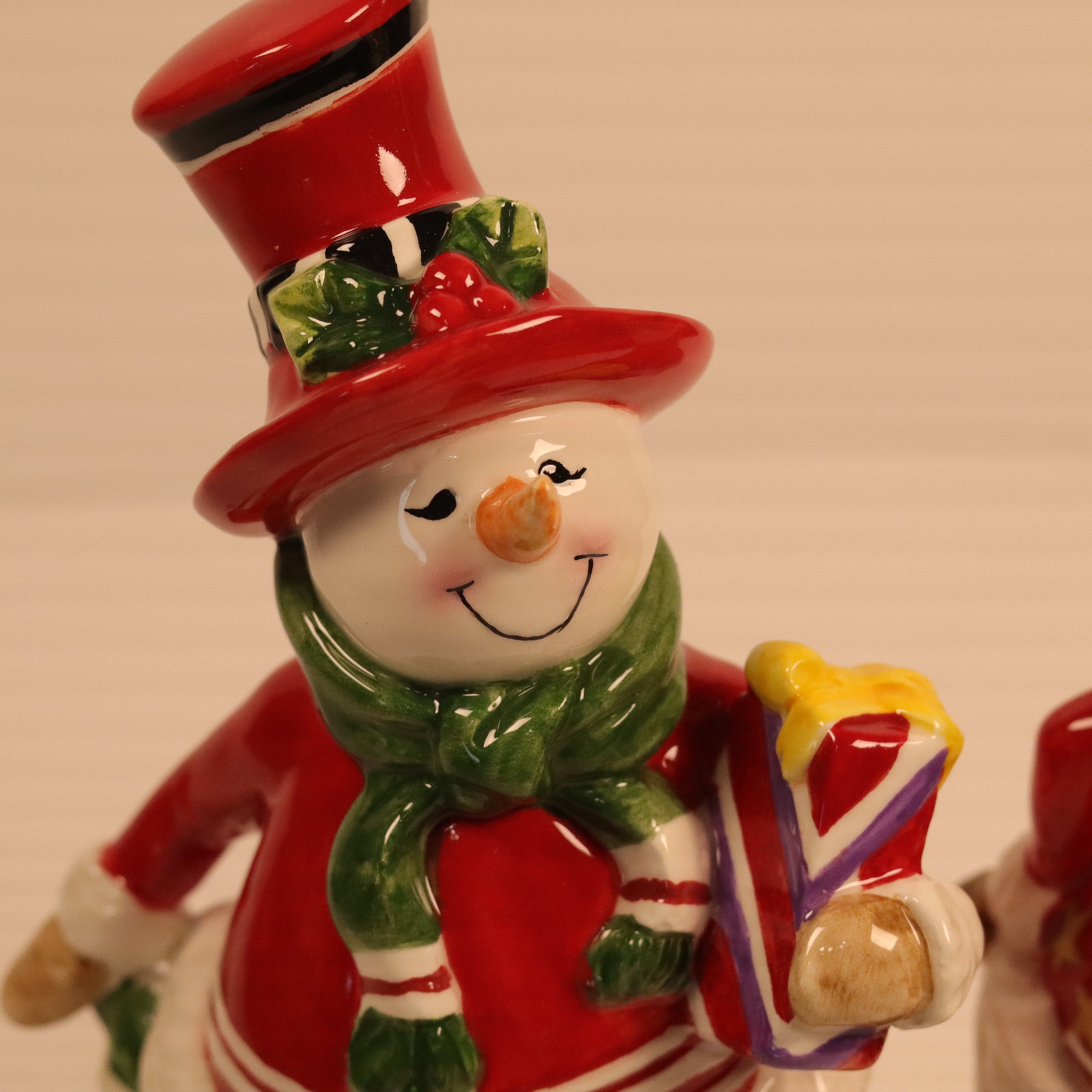 Snowman figurine with a red top hat, green scarf, and holding a colorful bag on a beige background.